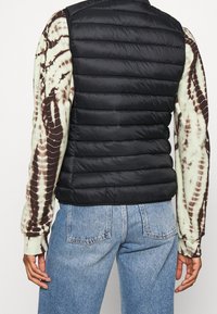 Sort puffer-vest med horisontalt quiltet design, båret over en tie-dye langærmet skjorte med grønne og brune farver, kombineret med lyse denimjeans.