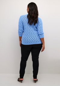 Himmelblauer Strickpullover mit einem Lochmuster und Dreiviertelärmeln, kombiniert mit schwarzen, eng anliegenden Hosen und schwarzen, spitzen High Heels.