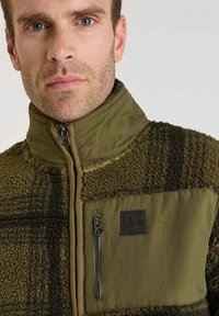 Groene fleecetrui met een ritssluiting bij de kraag, gestructureerde stof en een borstzak met ritssluiting. Logo-opdruk aan de linkerkant.