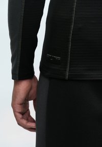 Čierne tričko s dlhým rukávom, vyrobené z textúrovaného materiálu, s viditeľným šitím a štítkom Nike Tech na dolnej časti chrbta. Úzky strih.