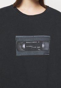 Musta huppari, jossa on kuva VHS-nauhasta ja teksti "MAISON MARGIELA AUTONME/HIVER 2020 PAL." Kangas näyttää pehmeältä ja siinä on rento istuvuus.