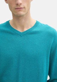 Pulover teal cu decolteu în V, realizat dintr-un material moale, având un mic logo pe piept și detalii ribbate la decolteu și manșete.