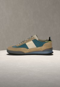 Αθλητικό παπούτσι casual με μπεζ σουέντ και τιρκουάζ υφασμάτινα πάνελ, λευκή κάθετη ρίγα στο πλάι, γκρι σόλα και πιστοποιημένη γλώσσα φτέρνας σε ουδέτερο φόντο.