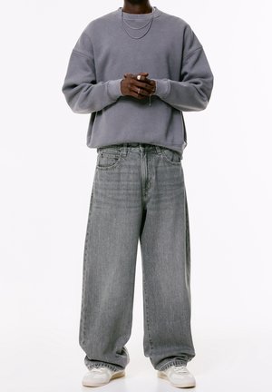 Personne portant un sweat-shirt gris oversize, un jean gris clair baggy, des baskets blanches, deux colliers en argent et des bagues, mains croisées devant.