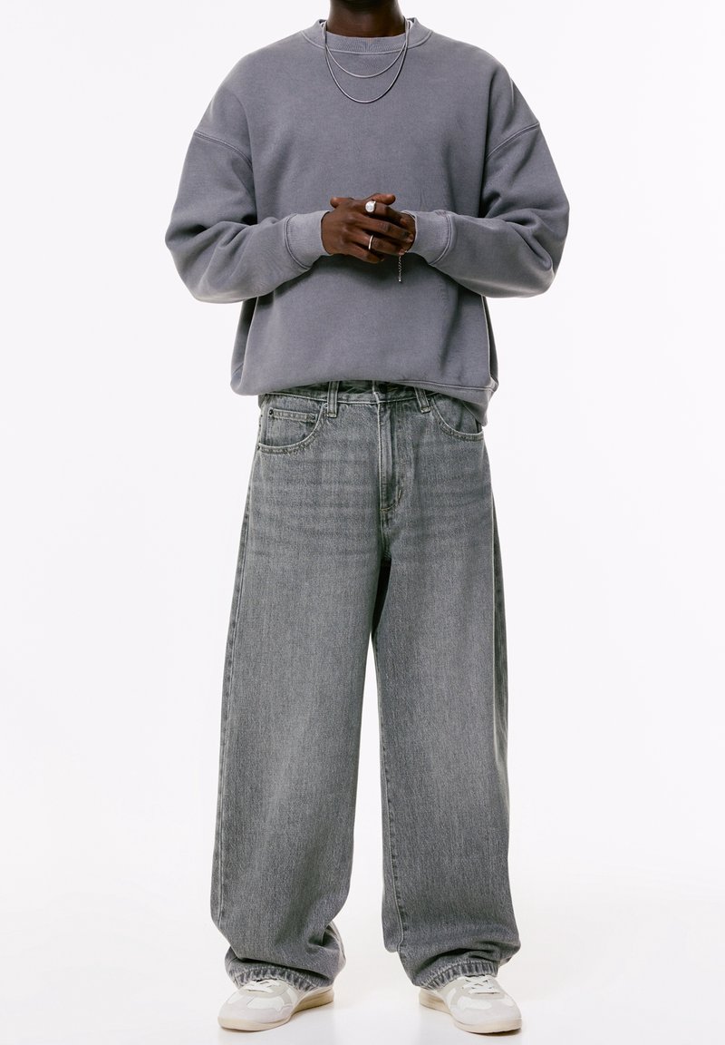 Personne portant un sweat-shirt gris oversize, un jean gris clair baggy, des baskets blanches, deux colliers en argent et des bagues, mains croisées devant.