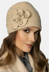 Chapeau en laine beige avec un design ajusté, présentant des accents de tissu roulé décoratifs sur le côté. Texture douce et finition lisse.