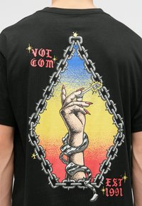 Zwarte T-shirt met een afbeelding van een geketende hand die een sigaret vasthoudt, omgeven door een driehoekige keten en een achtergrond met een vervaging van blauw naar geel.
