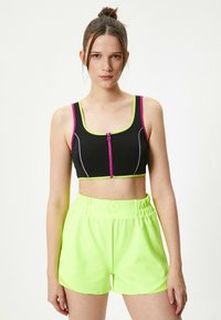 Sort crop top med lyserøde og grønne detaljer, der har en frontlynlås. Neon grønne shorts med elastisk talje og glat tekstur.