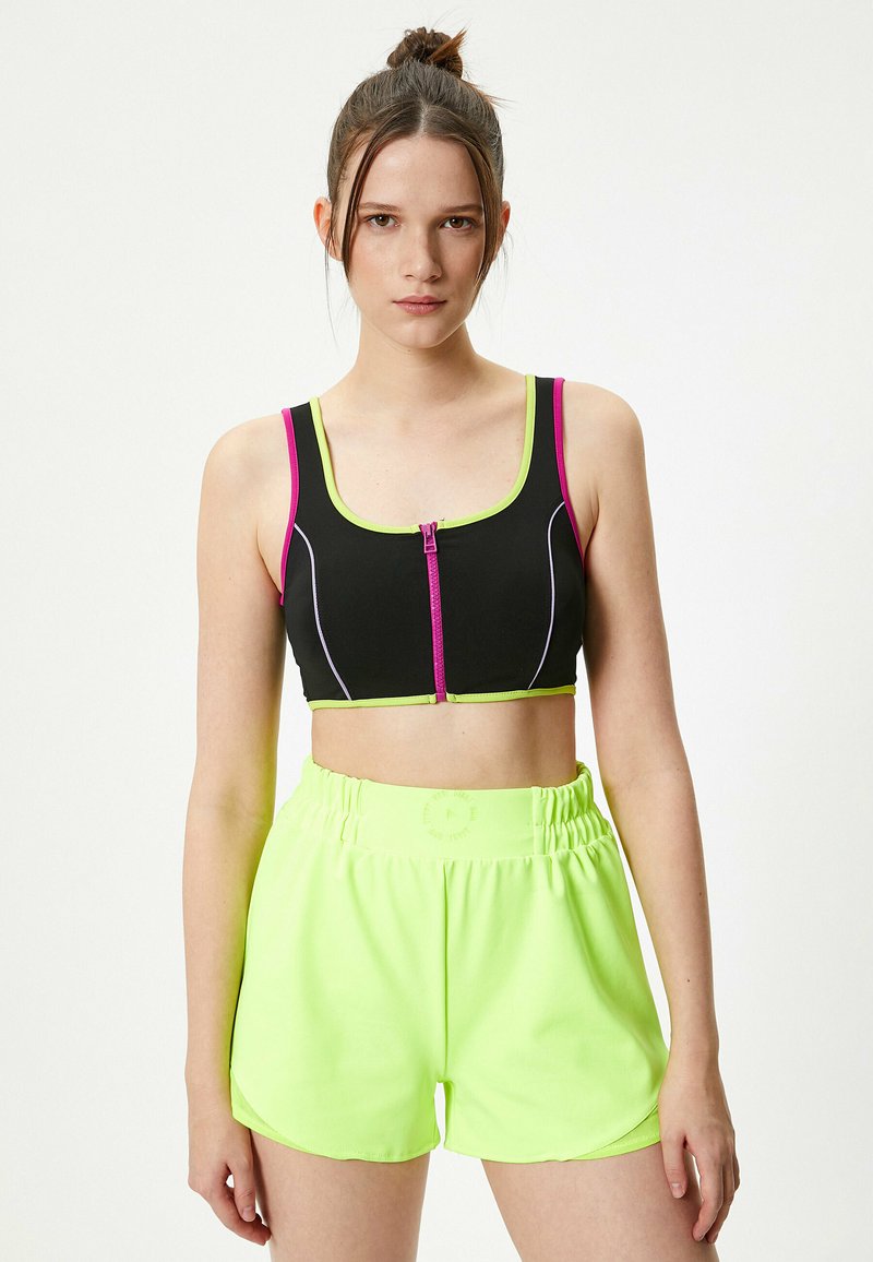 Sort crop top med lyserøde og grønne detaljer, der har en frontlynlås. Neon grønne shorts med elastisk talje og glat tekstur.