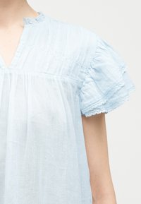 Γαλάζιο βαμβακερό πουκάμισο με ραφές πιερέ, λαιμόκοψη σε σχήμα V και κοντά στρωματοποιημένα μανίκια με βολάν και δαντέλα στο τελείωμα.