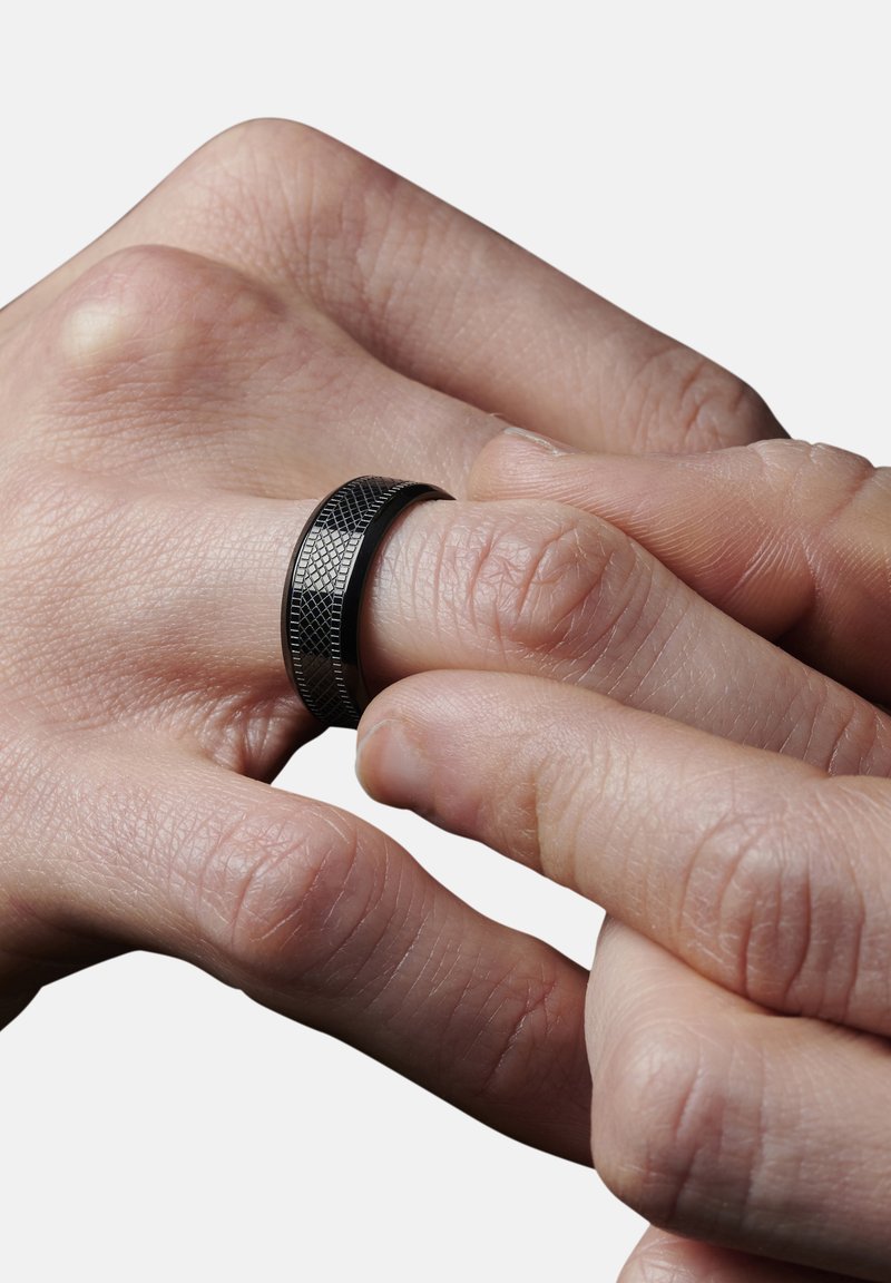 Sort metalring med et struktureret mønster, der sidder tætsluttende på en finger. Den glatte overflade og en let glans fremhæver designet.