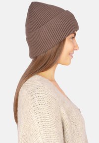 Braune Rippstrick-Beanie mit umgeschlagenem Bündchen, getragen von einem Modell. Die Textur ist weich mit vertikalen Linien und passt zu einem hellbeigen Pullover.