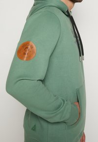 Grön hoodie i bomull med svart dragsko, en brun läderlapp på ärmen och en känguruficka. Strukturerad yta.