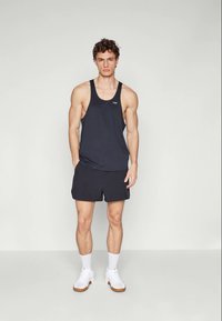 Jeune homme debout portant un débardeur de sport sans manches noir, un short noir, des chaussettes blanches et des baskets blanches devant un fond clair uni.