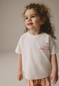 Camiseta blanca de algodón con el texto "Disney Princess" en naranja y un gráfico de corona. Combinada con pantalones a rayas en melocotón y blanco.