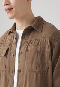 Camisa marrón de botones con dos bolsillos en el pecho, herrajes plateados y etiqueta de logo de Levi's. Hecha de un tejido texturizado con detalles de costura visibles.