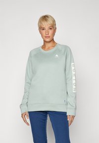 Sudadera verde clara hecha de tela suave, con un cuello redondo, puños acanalados y un logo en el pecho y la manga.