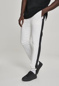 Hvite joggebukser med svart sideløsning, glidelåslomme og elastiske mansjetter, kombinert med svarte joggesko og hvite sokker.