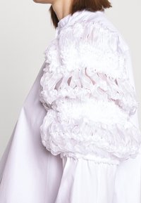 Witte blouse met puffed sleeves, versierd met geplooide, doorschijnende ruffles en een gladde body. De stof heeft een zachte textuur met een gestructureerd ontwerp.