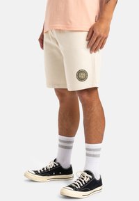Pantalones cortos de algodón beige con un parche redondo de logo caqui en la pierna izquierda. Combinados con zapatillas negras y calcetines blancos con rayas grises.