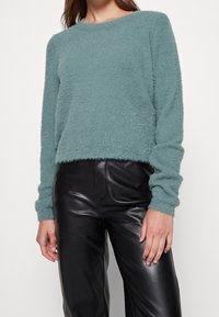 Camiseta de punto esponjosa verde menta con cuello redondo y mangas largas, combinada con pantalones de cuero negros de tiro alto, de textura suave.