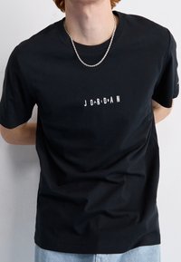 T-shirt di cotone nera con collo rotondo e maniche corte, con una grafica testuale consumata "JORDAN" in bianco. Jeans in denim azzurro chiaro visibili.
