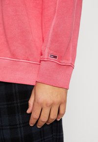 Sudadera de color coral con puños acanalados, que presenta una pequeña etiqueta de logo. Tela suave con una textura lisa y un corte holgado.