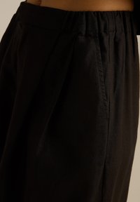 Czarne spodnie z elastycznym pasem, z plisami i g�ładką, matową teksturą. Zbliżenie ukazuje szczegóły tkaniny i szwy.