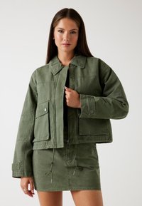 Jachetă scurtă de culoare verde oliv, cu guler ascuțit, buzunare frontale și capse argintii, asortată cu o fustă cu șnur în aceeași nuanță.