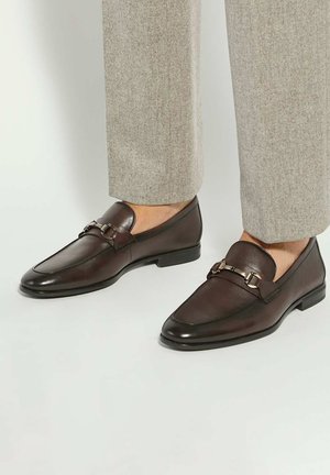 Brown leather loafers with a silver metal chain accent, rounded toe, and a low heel, paired with light gray tailored trousers.