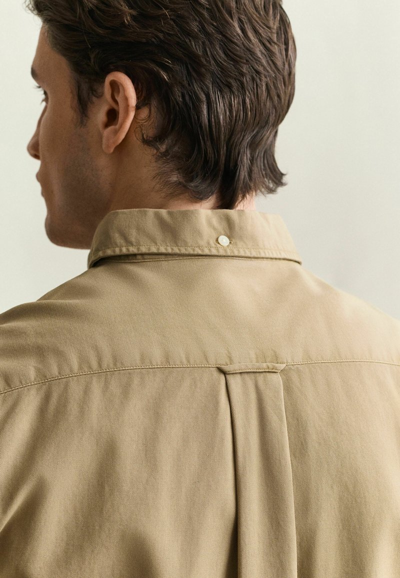 Man with short brown hair wearing a beige collared shirt, shown from the back and side against a neutral background.
