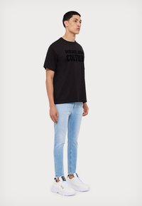 Schwarzes Baumwoll-T-Shirt mit strukturiertem Druck "VERSACE JEANS COUTURE". Kombiniert mit hellblauen Jeans und weißen Sneakers.