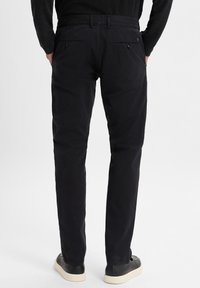 Schwarze Baumwollhosen mit schmalem Schnitt, die eine glatte Textur und aufgesetzte Taschen mit Knopfverschluss aufweisen, kombiniert mit schwarzen Sneakers.