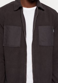 Giacca in pile grigio scuro con zip intera, due tasche sul petto e colletto a punta, indossata sopra una maglietta bianca.