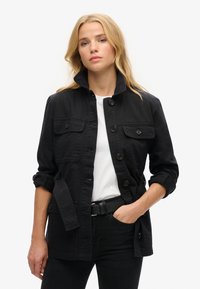 Chaqueta de algodón negra con un alto cuello, cierre de botones en el frente, dos bolsillos en el pecho y mangas enrolladas. Combinada con una camiseta blanca y un cinturón negro.