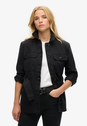 Schwarze Baumwolljacke mit hohem Kragen, vorderem Knopfverschluss, zwei Brusttaschen und hochgerollten Ärmeln. Kombiniert mit einem weißen T-Shirt und einem schwarzen Gürtel.