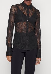 Chemisier en dentelle noire avec un design transparent, fermeture à boutons, col classique et poignets noirs unis. Détail de motifs en dentelle complexes.