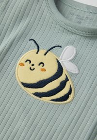 Vaaleanvihreä ribbikangas, jossa on keltainen brodeerattu mehiläinen, mustilla raidoilla, hymyilevä ilme ja valkoiset siivet. Tasainen pinta, pyöreä muoto.