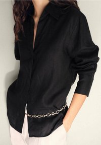 Chemise en lin noire avec un col boutonné, des manches longues et une coupe décontractée, accentuée par une ceinture en chaîne argentée.