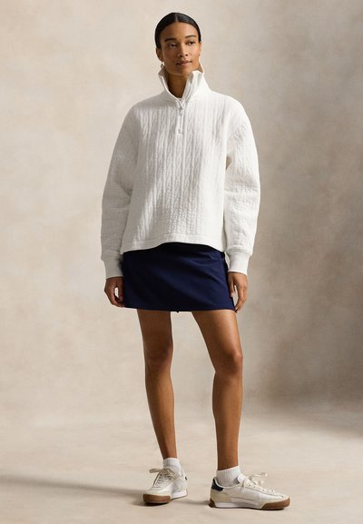 Λευκό πουλοβερί με καπιτονέ υφή και ψηλό γιακά, συνδυασμένο με σκούρο μπλε μίνι φούστα. Το μοντέλο φοράει λευκά αθλητικά παπούτσια με σόλες από καουτσούκ και λευκές κάλτσες.