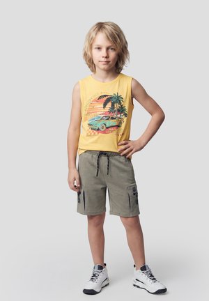 Niño rubio de pie con la mano en la cadera, vistiendo una camiseta sin mangas amarilla con diseño de coche y palmera, pantalones cortos cargo verdes y zapatillas blancas.