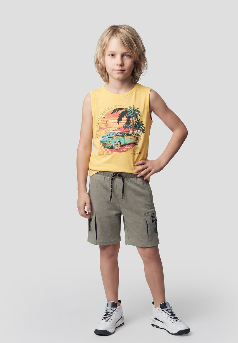 Blondes Kind steht mit der Hand in der Hüfte, trägt ein gelbes ärmelloses Shirt mit Auto- und Palmenmotiv, grüne Cargoshorts und weiße Turnschuhe.