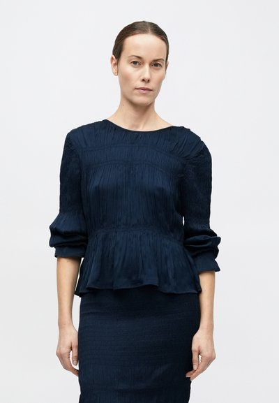 Femeie cu părul castaniu închis prins în spate poartă o bluză albastru închis plisată cu mâneci lungi și o fustă asortată, stând în fața unui fundal simplu și deschis la culoare.