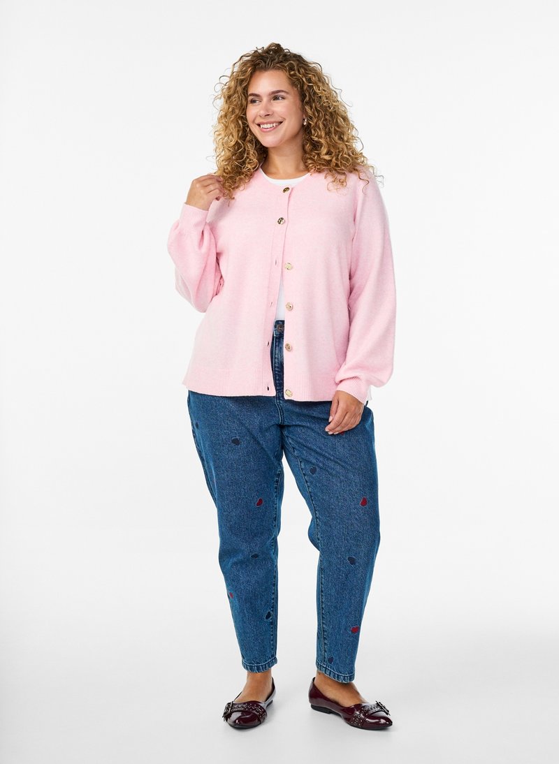 Vrouw glimlachend, draagt een lichtroze gebreide cardigan met knopen, wit hemd, blauwe jeans met hartjespatronen, en donkerrode platte schoenen, staand op een witte achtergrond.