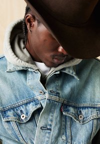 Šviesiai mėlyna džinsinė striukė su dviem krūtinės kišenėmis ir metaliniais mygtukais, dėvima ant pilkos gobtuvo, su tamsiai ruda platus kraštų skrybėle.