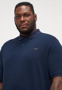 Námořnická polo tričko z bavlněné směsi s texturovaným povrchem, s knoflíkovou légou a malou značkovou nášivkou na hrudi.