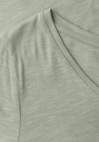 Gros plan sur le tissu vert clair d'un t-shirt à col en V, montrant la fine texture et les détails de couture sur l'épaule et le col.