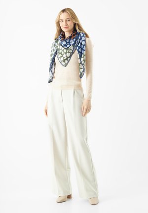 Écharpe en soie multicolore de forme triangulaire, ornée de motifs verts, bleus et blancs, associée à un pull beige et un pantalon blanc.
