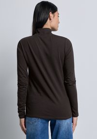 Vrouw met lang, donker haar draagt een donkerbruine, lange mouw turtleneck en een spijkerbroek, met haar rug naar de camera gekeerd tegen een effen lichte achtergrond.