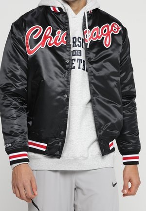 Zwarte satijnen varsityjack met "Chicago" in rode letters. Voorzien van gestreepte rode en witte geribbelde manchetten en tailleband. Draagt over een grijze hoodie.